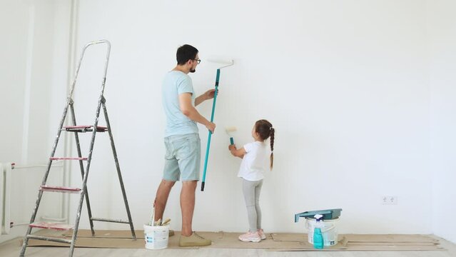 Happy attractive young father and cute little daughter makes repairs at home. Smiling girl and her daddy painting wall at living room.