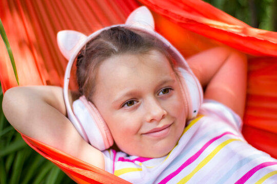 Cute little girl lies in a hammock and listening to music wearing wireless headphones at sunny summer day.