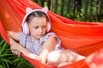 Cute little girl lies in a hammock and listening to music wearing wireless headphones at sunny summer day.