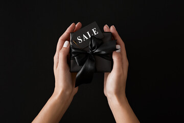 Top view photo of female hands holding black gift box with black satin ribbons and text on sale tag on isolated black background. Black Friday Sale template.