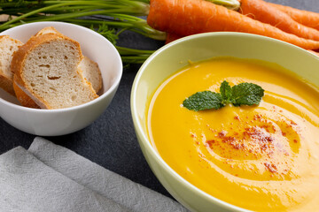 Horizontal image of bowl of carrot soup with garnish, raw carrots and sliced bread on slate