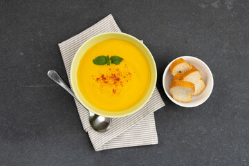 Horizontal image of bowl of carrot soup with garnish, napkin, spoon and bread on slate, copy space