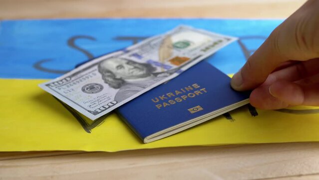 Hand Puts a Biometric Passport of Ukraine with Cash on a Background of Flag. Drawing of yellow, blue Ukrainian flag with title stop war lie on table. Money, bribe. War, migrants leaving the country.