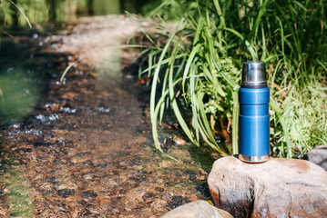 Vacuum camping thermos on stone near stream in forest on sunny day, copy space. Equipment for hiking, camping