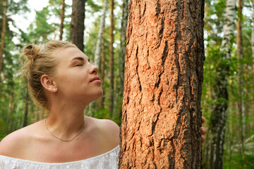A young woman stands near a tree with her eyes closed and inhales the smell of the forest.