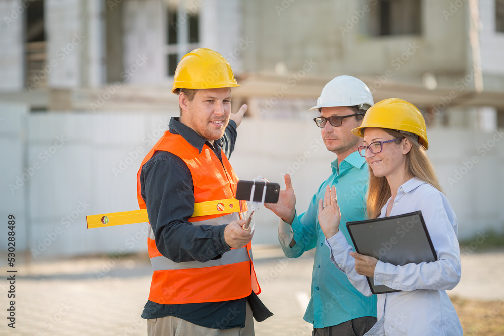 Wall mural men and a woman in yellow and white helmets, blue shirts are standing at a construction site with do