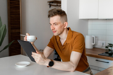 Leisure time. Young man reading news on digital tablet or surfing internet and enjoying morning coffee, sitting in kitchen interior at home