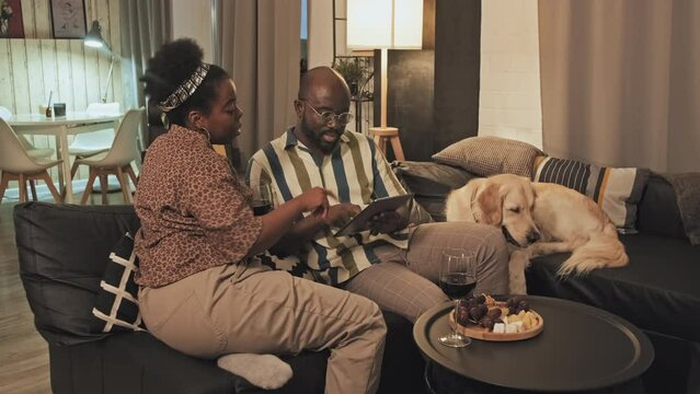 Medium long of young African American hetero couple with dog sitting on couch in living room, talking, using tablet computer