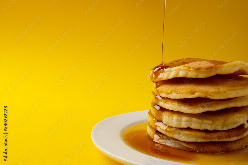 Poster horizontal image of maple syrup pouring onto stack of pancakes, yellow background and copy space