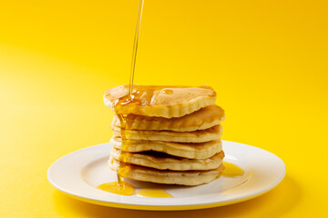 Horizontal image of maple syrup pouring onto stack of pancakes, yellow background and copy space