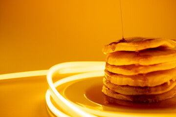 Horizontal image of maple syrup pouring onto stack of pancakes, yellow background and copy space