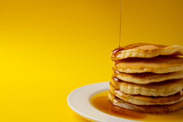 Horizontal image of maple syrup pouring onto stack of pancakes, yellow background and copy space