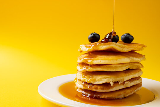 Horizontal image of maple syrup pouring onto pancakes and blueberries, on yellow with copy space