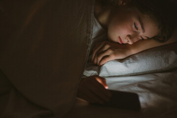 child teenager girl lies in bed in evening or at night and looks into phone. mobile addiction.