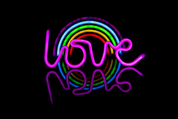 Image of vibrant neon love text and glow sticks forming rainbow over black background and copy space