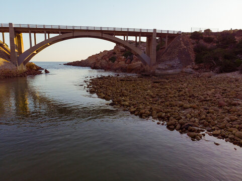 Photo of bridge over water with pebbles on shore and blue sky