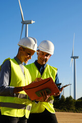 Two engineers checking the progress of wind turbines while working in eolic park.