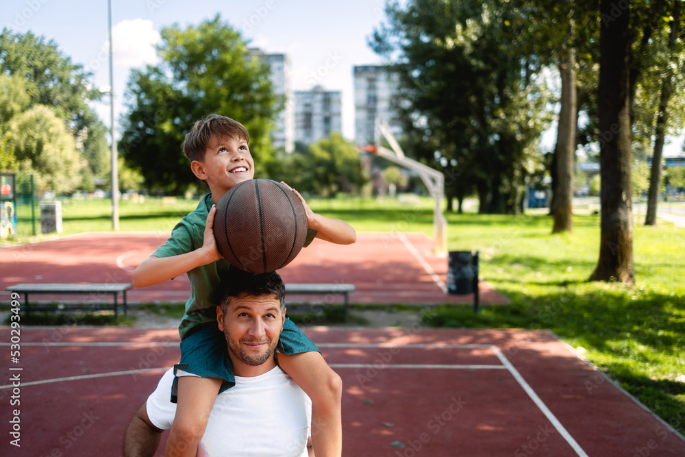 Wall mural the father holds his son on his shoulders, helps him throw the ball into the basket. they wear casua