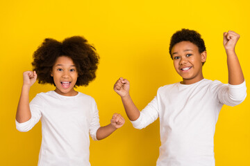 Portrait of two excited successful pupils raise fists celebrate triumph isolated on yellow color...