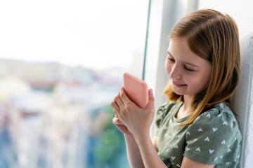 Pretty preteen girl looking at smartphone and smiling at home close to window with daylight....