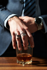 Cropped close-up shot of a man in a suit holding a glass of whiskey over the table in a dark room. A man's hand is with two snake rings. Front view.