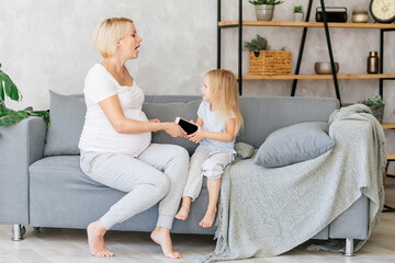 A pregnant young girl is sitting on the sofa with her little daughter and playing on the phones. The concept of the problem of addiction to gadgets in children and adults