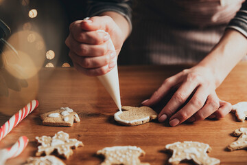Christmas, New Year food preparation. Xmas gingerbread cooking, making and decorating freshly baked cookies. Closeup on cropped shot with woman hands holding icing and mastic, decorating cookies