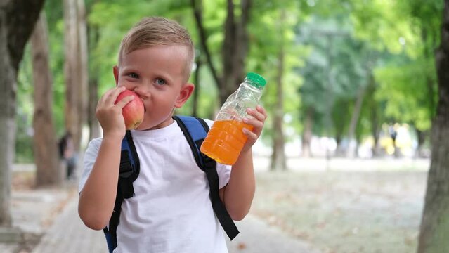 Little boy eats an apple and drinks juice. A child with a backpack has a snack before school.