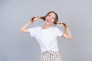 Fototapeta na wymiar Cheerful lady in striped pants fooling around in studio and holding her hair with her hands and laughing. Charming girl in white t-shirt is dancing on gray background. Caucasian girl counts her teeth