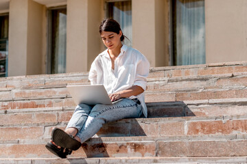 A young dark-haired woman in a white shirt and jeans uses a laptop sitting on the steps on a city street on a summer day.Remote work, freelance,online education.