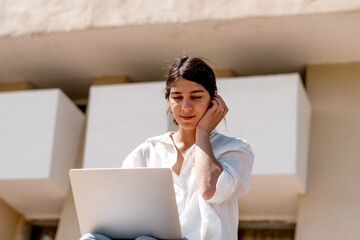 A young dark-haired woman in a white shirt and jeans uses a laptop sitting on the steps on a city...