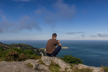 Fototapeta na wymiar Young man sits on top of a rocky hill in Killiney and watches the sea on a gorgeous sunny day, Killiney, Ireland