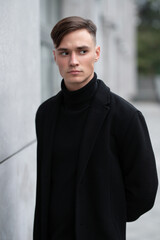 Fashion portrait of a young guy in black clothes of model appearance on the street among skyscrapers and alleys