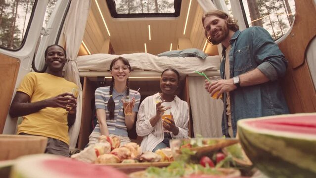 Portrait of delighted young men and women getting together in cozy house on wheels parked at campsite, smiling at camera with drinks in plastic cups
