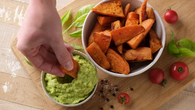 Dipping baked sweet potato wedge into vegan avocado sauce. Healthy food, vegetables for dinner or snack