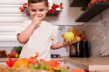 Smiling cute boy standing at counter with fruits, autumn leaves and pumpkin. Autumn decorated kitchen. Holidays concept.