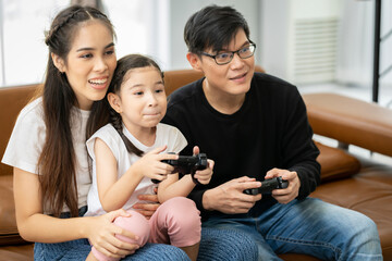 Family of Asian parents and daughters Spend your holidays together playing video games on the sofa in your house with fun and happy smiles.