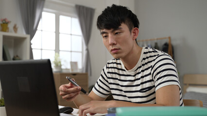 closeup asian Korean university boy holding face with boredom is looking away into empty area and spinning pen while learning from home watching online video lecture.