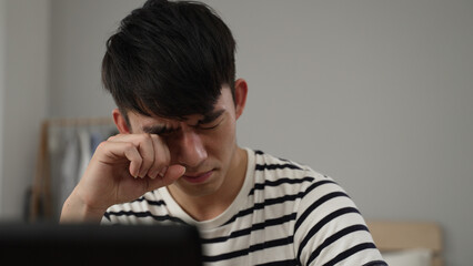 closeup view of an overworked Korean young man having sudden blurry vision is rubbing his eyes while working from home on the computer.