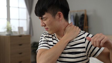 closeup shot asian Chinese male suffering shoulder stiffness is having a massage with a grimacing face while learning from home in the bedroom