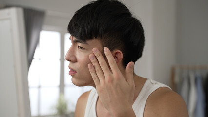 Fototapeta na wymiar frustrated asian Japanese guy looking at mirror is touching his face feeling unhappy about first wrinkles while going through daily skincare beauty routine at home.