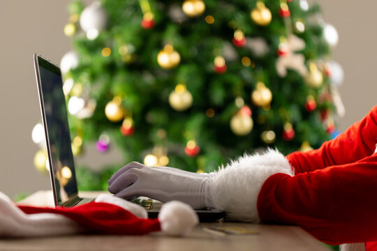 Image of hands of santa claus using laptop with christmas tree in background