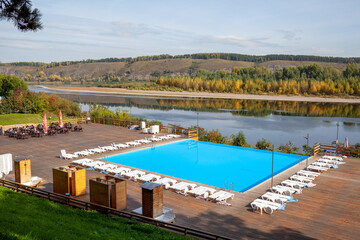 Fototapeta na wymiar Blue pool without people on the riverbank in autumn. A great place to relax and unwind in nature. The pool and sun beds are waiting for visitors