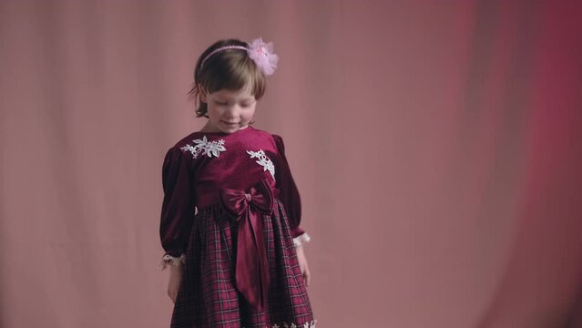 Cute little girl in an vintage burgundy dress is dancing, rejoicing and making funny faces. The concept of children theater productions and developing acting circles. Pink background