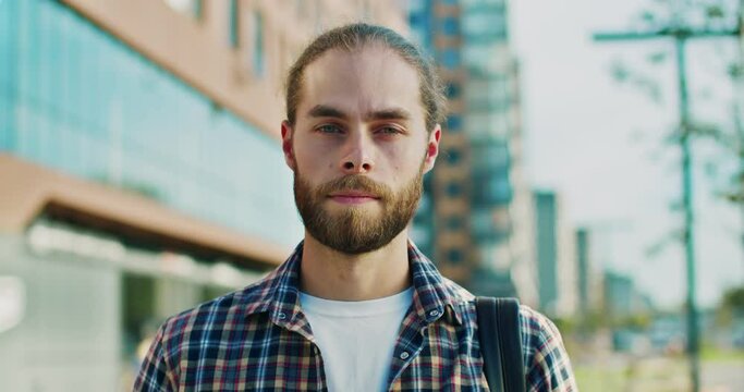 Portrait of young Caucasian handsome man with beard and hair in tail. Buildings on background. Outside. Attractive male with backpack and in motley shirt looking to camera at street. Close up.