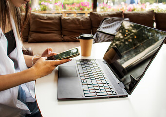 working on a laptop close-up, female hands hold a smartphone while working on a laptop, a glass of coffee is nearby on the table