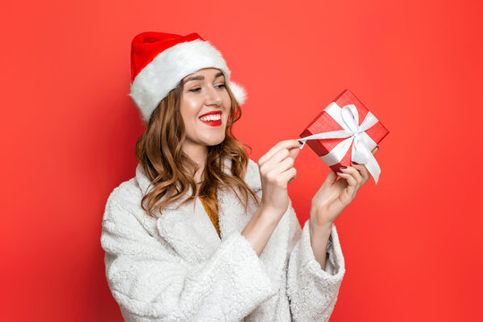 Happy cute young woman in christmas holiday hat smiling and opening red gift box with white ribbon isolated over red studio background