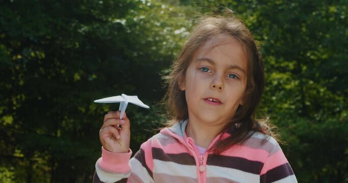 Cute little girl with a paper plane in the park. Child launches a paper airplane close-up. Cute little girl has fun playing with a toy paper plane. Concept of outdoor activities in summer.