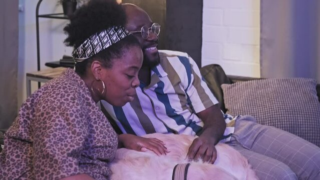 Medium Long Side View Of African American Family Couple Relaxing On Couch, Watching TV In Living Room At Night, Talking And Smiling, Petting Sleeping Dog