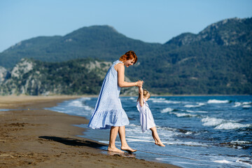 Cute little toddler girl and mother in blue dresses runnig and playing with waves on the wild beach. Turkey, Iztuzu beach,  Dalyan. Mom and daughter